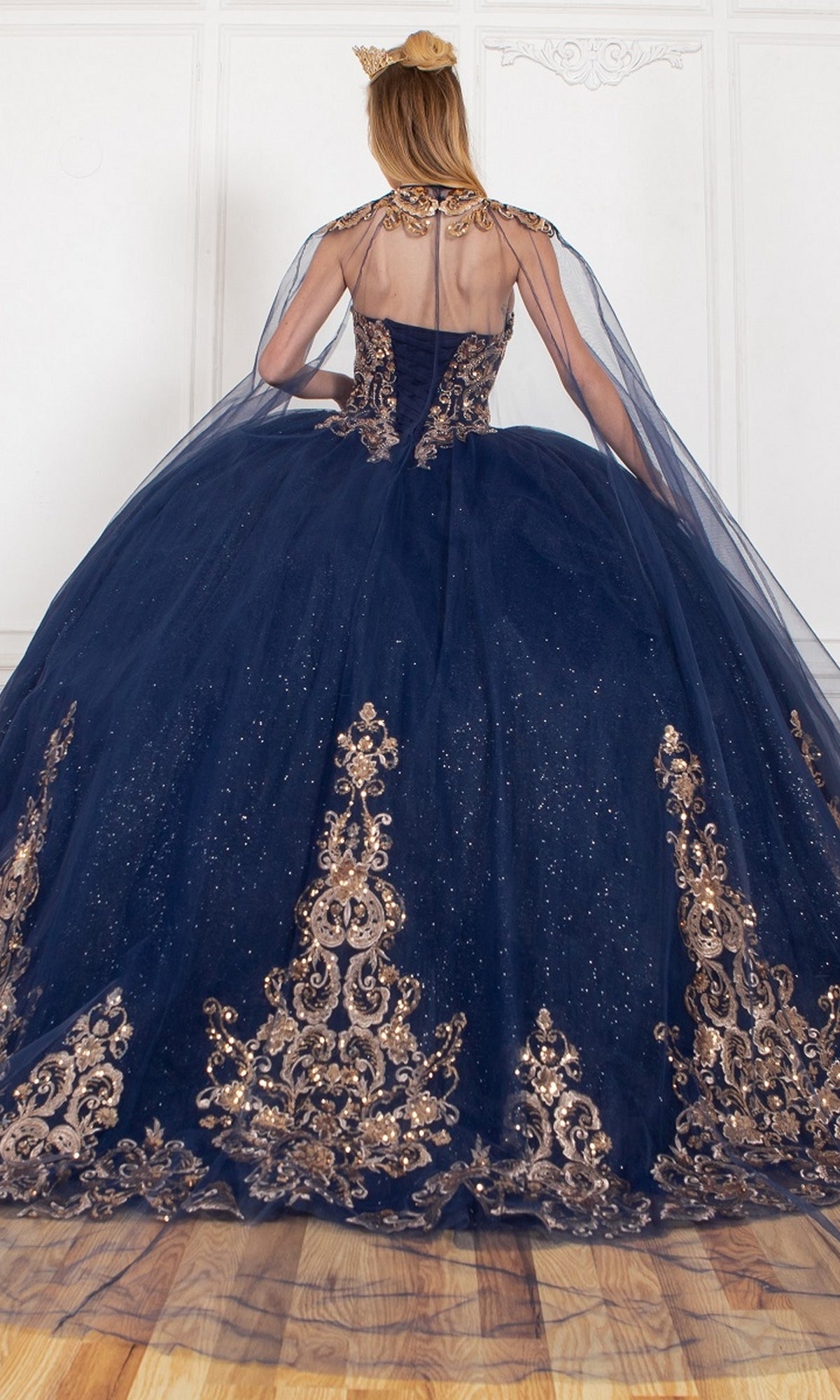 A person stands facing away in a white room, wearing the Quinceanera Ball Gown 8063J: a navy blue ball gown with gold embroidery, strapless sweetheart neckline, and a sheer glitter tulle cape.